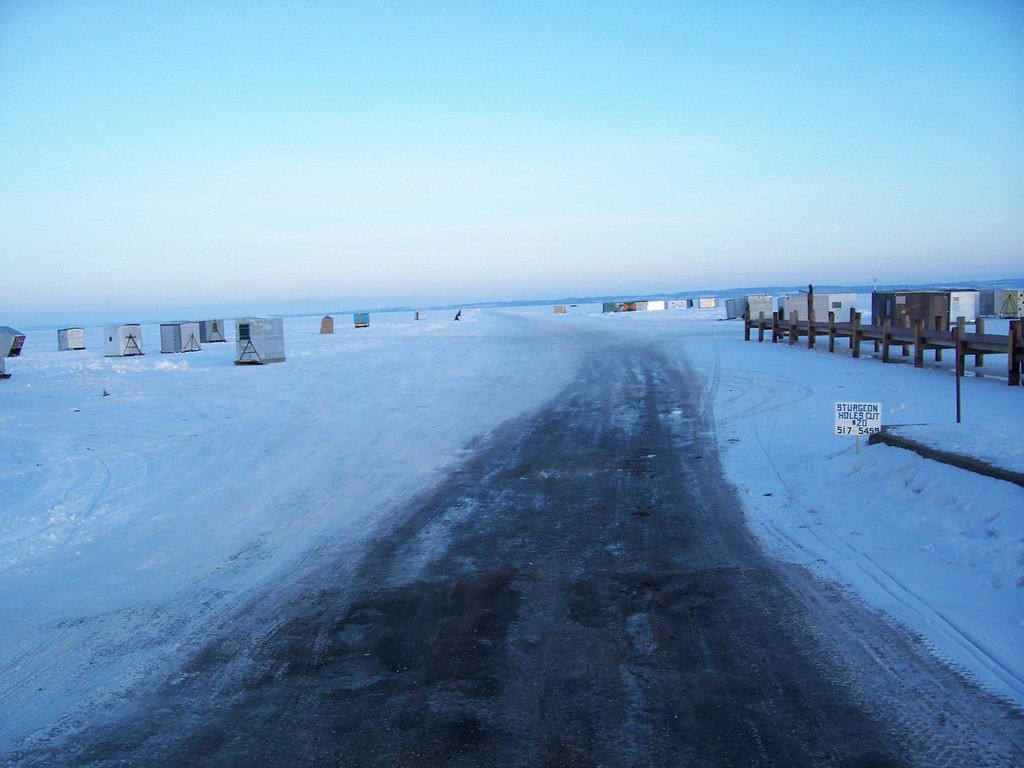 A plowed ice road running out onto frozen Lake Winnebago, lines of ice shanties on either side and a sturgeon-season sign at the roadside